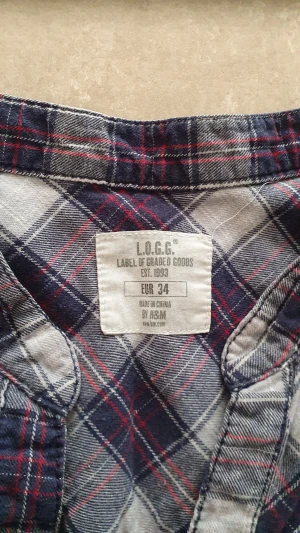 Skjortklänning - Rutig skjortklänning från H&M i blått, vitt och rött. Klänningen har trekvartsärmar med knappdetaljer, v-ringad hals och knappar framtill. Volangkant nedtill ger en chill vibe. Perfekt för dig som gillar avslappnad stil med lite edge.