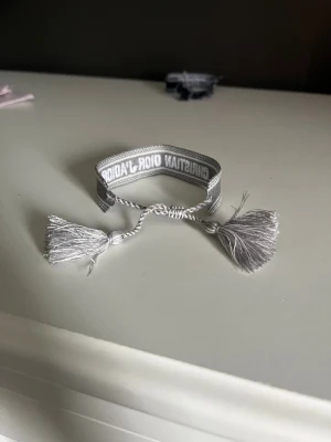 Grått Christian Dior Armband - Snyggt grått friendship armband från Christian Dior med broderad logga och två dekorativa tofsar. Armbandet har en justerbar knytning och är gjort i ett mjukt textilmaterial som känns bekvämt på handleden. Perfekt för att ge din outfit en lyxig touch.