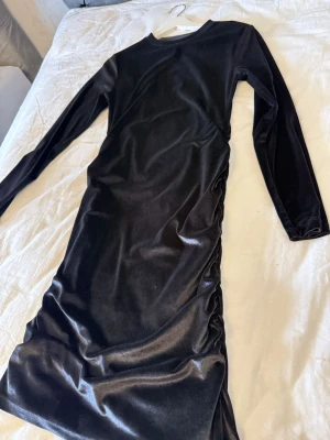 Svart långärmad sammetsklänning - Snygg svart klänning i mjuk sammet med lång ärm och rund hals. Klänningen har en figurnära passform och är helsvart utan mönster, perfekt för dig som gillar stilrena plagg med lite lyxig känsla. Längden är lång och materialet ger en cool glans.