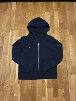 Mörkblå hoodie från GANT med dragkedja - Snygg mörkblå hoodie från GANT med hel dragkedja i metall och huva med snörning. Tröjan har ribbade muddar, fickor framtill och broderad GANT-logga på bröstet. Perfekt för chill dagar och enkel att matcha med jeans eller joggers. Jag skulle säga att passar bådd i M och S storleken.