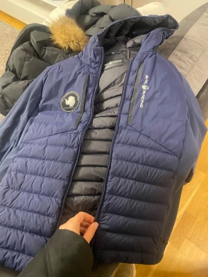 Blå Sail Racing pufferjacka med huva - Säljer en mörkblå pufferjacka från Sail Racing med snyggt broderat märke på ärmen och logga på bröstet. Jackan har huva med pälsdetalj, dragkedja framtill och quiltad design. Perfekt för kalla dagar och har en clean, sportig look.