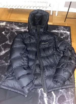 Svart pufferjacka från Peak Performance - Säljer en svart pufferjacka från Peak Performance med hög krage och huva. Jackan har en tydlig dragkedja framtill och är quiltad för extra värme. Perfekt för kalla dagar och har en stilren look som funkar till det mesta.