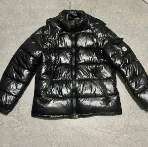 Svart pufferjacka från Moncler - PRIS KAN DISKUTERAS LÅGT//Svart glansig pufferjacka från Moncler med klassisk logga på ärmen. Jackan har hög krage, två sidofickor med dragkedja och quiltad design. Materialet är syntet med dunfyllning, perfekt för kalla dagar. Stängs med dragkedja framtill och har coola detaljer inuti.