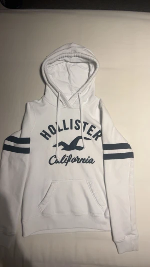Vit hollister hoodie - Vit hoodie från Hollister i storlek XS med marinblå tryck och detaljer. Tröjan har huva med snörning, känguruficka och två marinblå ränder på ärmarna. Mjuk insida i fleece och klassisk Hollister-logga framtill. Perfekt för chill dagar.