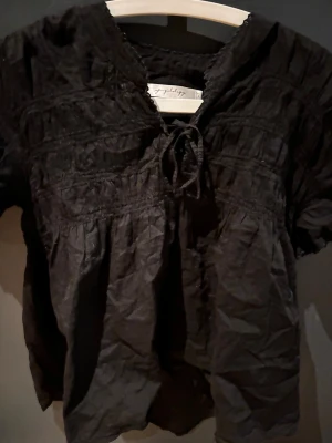 Svart blus med knyt från Joyfulday - Säljer en svart blus med korta ärmar och snygg knytning vid halsen. Blusen har ett lätt rynkat tyg och en luftig passform som ger en cool och avslappnad vibe. Perfekt för dig som gillar stilrena och bekväma plagg.Passat storlek M också 