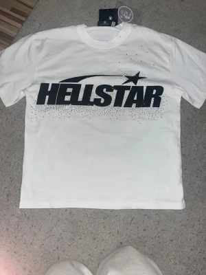 Vit Hellstar t-shirt med tryck - Säljer en vit Hellstar t-shirt i 100% bomull med stor svart logotyp och stjärntryck på framsidan samt coolt grafiskt motiv på ryggen. T-shirten har rund hals och korta ärmar, perfekt för dig som gillar streetwear och snygga prints.