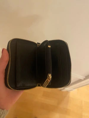 Svart plånbok i skinn med dragkedja - Snygg svart plånbok i skinn med guldfärgad dragkedja runtom. Plånboken har flera fack inuti och ett separat myntfack med dragkedja. Stilren och kompakt design som passar perfekt i väskan eller fickan.