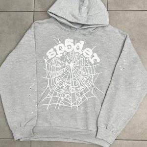 Grå hoodie från Sp5der med tryck - Säljer en grå hoodie från Sp5der med stort vitt spindelnäts-tryck och logga på framsidan. Tröjan har huva, ribbade muddar och lång ärm. Perfekt för dig som gillar streetwear och coola detaljer.