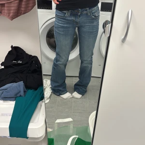 Blå bootcut jeans med fickdetaljer - Snygga blå jeans med bootcut passform och klassisk femficksdesign. Jeansen har coola bakfickor med lock och knapp, samt lätt slitna detaljer framtill och baktill. Perfekta för dig som gillar en avslappnad men ändå trendig look. Storlek S