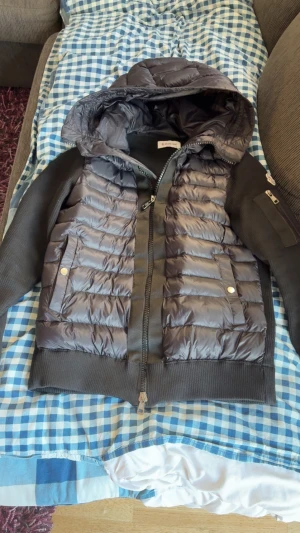 Svart pufferjacka från Moncler - Svart pufferjacka från Moncler med huva och ribbade muddar. Jackan har quiltad framsida i glansigt material, dragkedja och fickor med tryckknappar. Ärmarna är i stickad kvalitet och har en ficka med dragkedja på ena armen. Perfekt för kyliga dagar.