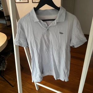 Ljusgrå pikétröja med tigerbrodyr - Snygg ljusgrå pikétröja från Tiger of Sweden med klassisk krage och korta ärmar. På bröstet finns en broderad tiger i svart och vitt som ger en cool detalj. Tröjan har knappar vid halsen och är gjord i mjuk bomull som känns skön mot huden.