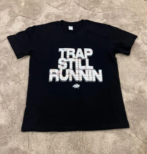 Svart t-shirt med tryck Trap Still Runnin - Svart t-shirt från Gildan med stort tryck på bröstet där det står 'Trap Still Runnin' i vit och grå 3D-stil. Klassisk rund hals och korta ärmar, perfekt för dig som gillar streetwear och statement-plagg.