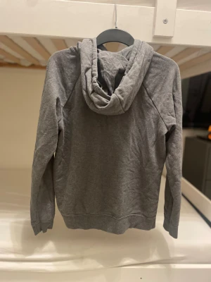 Grå hoodie från Nike med huva - Grå hoodie från Nike med vit logga på bröstet och vita snören i huvan. Tröjan har en stor magficka och är långärmad. Mjuk och skön bomullsmix som passar perfekt när du vill chilla eller dra till gymmet. Klassisk och enkel stil med normal passform. 