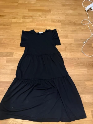 Svart långklänning med volanger - Säljer en svart långklänning med rund hals och korta ärmar. Klänningen har en avslappnad passform och volangdetaljer i två lager som ger en snygg och flowy siluett. Perfekt för dig som gillar enkel och stilren design.