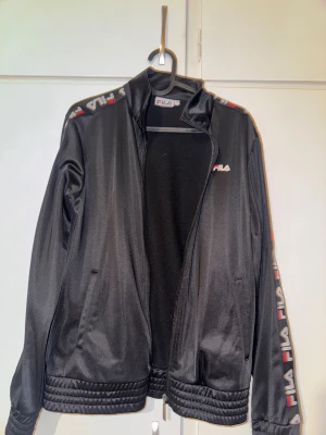Svart FILA kofta  - Svart vindjacka från FILA i storlek S. Jackan har dragkedja framtill, ribbade muddar och FILA-logga på bröstet samt längs ärmarna. Materialet är polyester. Sparsamt använd