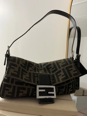 Fendi axelväska med FF-mönster - Snygg Fendi axelväska med ikoniskt FF-mönster i mörkbrunt och svart. Väskan har en rektangulär form, justerbar axelrem i svart skinn och en stor silverfärgad spänne framtill. Materialet är tålig canvas med detaljer i skinn och dragkedja på insidan.