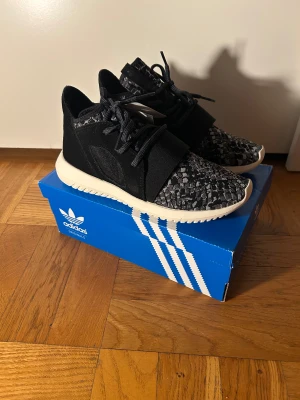 Svarta Adidas sneakers med mönster - Säljer ett par svarta Adidas sneakers med grått och svart mönster på ovandelen. Skorna har snörning, platt vit sula och rund tå. Materialet är syntet och textil, vilket gör dem riktigt sköna och lätta. Perfekt för dig som gillar stilrena och sportiga sneakers. Strl 38, 38.5 och 42 finns i lagret.