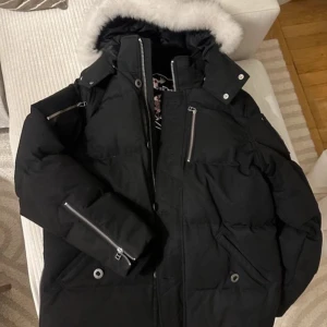 Svart Moose Knuckles dunjacka med päls - Svart dunjacka från Moose Knuckles med fluffig vit päls på huvan, flera dragkedjor och snygga metalldetaljer på ärmen. Jackan har lång ärm, hög krage och praktiska fickor. Perfekt för kalla dagar och har en cool streetstil. !ÄKTA FÅTAL ANVÄND!
