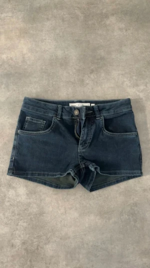 Mörkblå denimshorts med låg midja - Snygga mörkblå denimshorts med låg midja och klassisk femficksdesign. De har knapp och dragkedja framtill samt bälteshällor. Perfekta för varma dagar och en avslappnad stil.