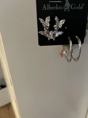 Silverörhängen fjäril & ring Albrekts Guld - Säljer ett set med silverfärgade örhängen från Albrekts Guld. Ena paret är formade som fjärilar med glittrande stenar och det andra paret är klassiska släta ringar. Perfekt för dig som vill variera din stil med både detaljer och enkelhet.