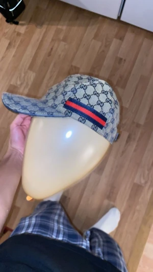 Keps - Säljer en klassisk keps i beige och blått. Kepsen har en böjd skärm och en snygg röd-blå rand på sidan. Perfekt accessoar för att lyfta din outfit med lyxig streetstyle.