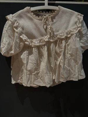 Vit blus med volang från Zara - Supersöt vit blus från Zara med puffiga korta ärmar och volangdetaljer längs axlarna. Ovanpartiet är stickat med knytning fram och spetskant, medan resten är i mjuk bomull. Perfekt för dig som gillar romantisk och vintage-inspirerad stil.