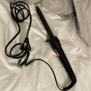 Locktång Svart - En svart locktång med konisk stav och lång sladd. Utrustad med en röd strömbrytare. Produkten ser ut att vara i gott skick utan synliga skador eller slitage. Perfekt för att skapa vackra lockar snabbt och enkelt.