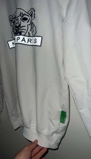 Vit sweatshirt från Kenzo med tiger - Säljer en vit sweatshirt från Kenzo i storlek M med det ikoniska tigermotivet och 'Kenzo Paris' broderat på bröstet. Tröjan har ribbade muddar vid hals, ärmslut och nederkant samt en grön Kenzo-tag på sidan. Perfekt för dig som gillar streetwear och snygga loggor.