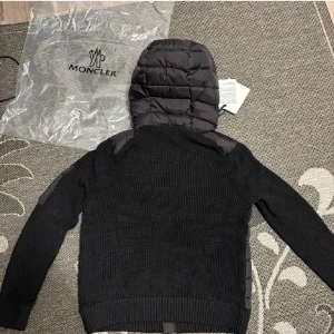 Svart Moncler pufferjacka med huva - Svart pufferjacka från Moncler med stickade ärmar och quiltad kropp. Jackan har huva, dragkedja framtill och Moncler-logga på ärmen. Materialmix av polyester och stickad textil, samt praktiska fickor med knappstängning och en extra ficka med dragkedja på ärmen.