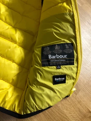 Svart och gul dunväst från Barbour - Snygg dunväst från Barbour International i svart med gul insida och gul dragkedja. Västen är quiltad och har Barbour-logga på bröstet. Perfekt för lager-på-lager och ger en cool kontrast med den färgstarka insidan.