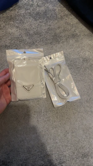 Vit plånbok korthållare runt nacken  - Supersnygg vit plånbok från Prada med klassisk trekantig logga i silver på framsidan. Kommer med matchande vit rem som enkelt fästs med metallspänne. Perfekt för kort och småsaker, stilren och minimalistisk design i skinn.