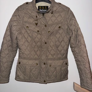 Beige quiltad jacka från Barbour köpt på Pondus - Säljer en beige quiltad jacka från Barbour med snygga detaljer som knappar och hög krage. Jackan har flera fickor framtill och ett klassiskt rutmönster. Perfekt för dig som gillar stilren och tidlös design.