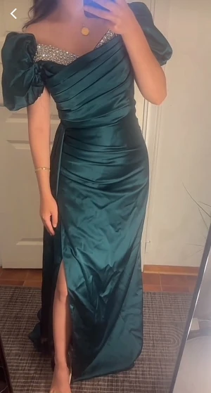 Mörkgrön långklänning med puffärm - Säljer en elegant mörkgrön långklänning med glittrande stenar vid halsringningen och puffiga korta ärmar. Klänningen har omlott-detaljer upptill, slits framtill och är gjord i ett glansigt satinliknande material som ger en lyxig känsla. Perfekt för bal eller fest!