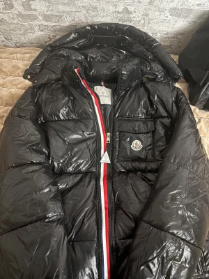 Moncler Montbeliard hooded down jacket - Svart pufferjacka från Moncler med glansig finish, huva och ikonisk logga på bröstfickan. Jackan har dragkedja med röd och vit kant, samt innerficka med serietidningsmönster. Perfekt för dig som vill ha en snygg och varm jacka med street vibe.