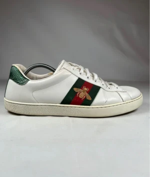 Gucci Ace Beatel - Gucci Ace sneakers i vitt skinn med klassiska gröna och röda ränder på sidan och broderad guldfärgad bi. Grön och röd metallic-detalj på hälen, platt sula och rund tå. Snygg snörning framtill och lyxig känsla som sticker ut.