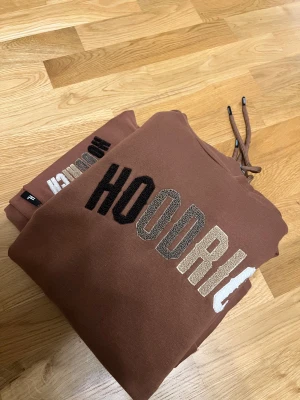 Brun HOODRICH hoodie med broderi - Säljer en brun hoodie från HOODRICH med broderad logga i svart, beige och vitt på bröstet. Hoodien har huva med snörning och är tillverkad i mjuk bomull med fleece på insidan. Perfekt för chill dagar och streetwear-stil.
