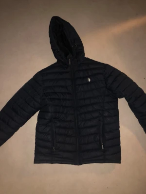 Svart pufferjacka från U.S. Polo Assn. - Svart pufferjacka från U.S. Polo Assn. med huva och dragkedja framtill. Jackan har quiltad design, två fickor med dragkedja och en liten logga på bröstet. Perfekt för kalla dagar och har en clean, sportig look. Den är i perfekt skick förutom att högra fickan är lite tröja o öppna/stänga men går säker o fixa det enkelt.