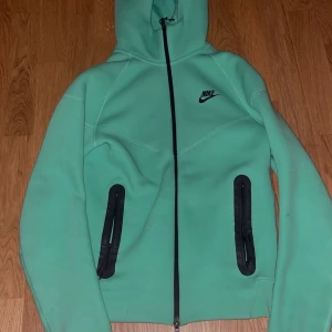 Mintgrön Nike hoodie med dragkedja - Fräsch mintgrön hoodie från Nike med svart logga på bröstet. Tröjan har hel dragkedja framtill, två svarta fickor med dragkedja och en skön huva. Materialet är mjukt och känns som polyester, perfekt för chill eller träning. Långärmad och snygg passform.storlek xs men passar S
