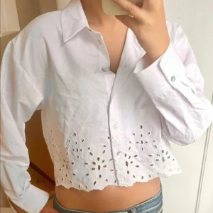 Vit croppad skjorta med broderi - Säljer en vit croppad skjorta med snygga broderade detaljer och hålmönster längst ner