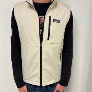 Beige väst från Patagonia med dragkedja - Snygg beige väst från Patagonia med svart dragkedja framtill och på fickorna. Västen har en stilren look med hög krage och logga på bröstet. Perfekt att slänga över en hoodie eller tröja för extra lager. Materialet känns lätt och smidigt. L passar även M!