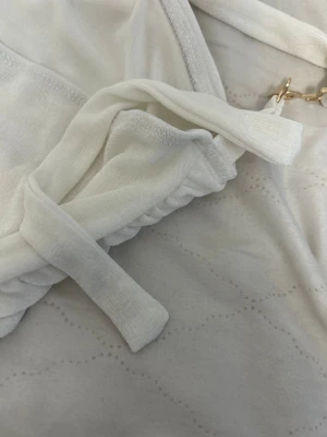 Vit oneshoulder topp med gulddetalj - Snygg vit oneshoulder topp med guldfärgad metallring på axelbandet. Toppen har en croppad passform och är gjord i ett mjukt, glansigt syntetmaterial med draperade detaljer. Perfekt för dig som gillar stilrena och trendiga plagg med en twist.