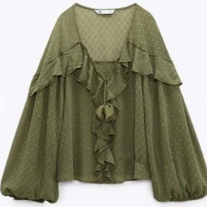 Olivgrön blus med volanger från Zara - Säljer en olivgrön blus från Zara med volanger och knyt framtill. Blusen har långärmade, luftiga ärmar och ett diskret prickigt mönster. Materialet är lätt och flowy, perfekt för dig som gillar boho-vibe och detaljer som ger extra stil.