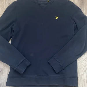 Svart sweatshirt från Lyle & Scott - Svart sweatshirt från Lyle & Scott med klassisk gul fågel-logga på bröstet. Tröjan har rund hals, ribbade muddar vid ärmslut och nederkant samt långärmad passform. Perfekt för dig som gillar stilren och enkel design. Kan kanske vara lite tight 
