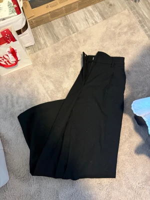 Svarta vida kostymbyxor - Säljer ett par svarta kostymbyxor med riktigt breda ben och hög midja. Byxorna har hällor för bälte och stängs med dragkedja och knapp. Perfekta för dig som gillar en stilren och trendig look.