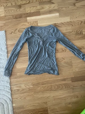 Grå långärmad blus från Intimissimi - Supersnygg grå långärmad blus från Intimissimi med v-ringad spetsdetalj både vid halsen och ärmsluten. Mjuk och tunn kvalitet med inslag av kashmir som gör den extra skön. Perfekt för dig som gillar stilrena och feminina plagg.