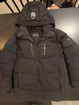 Svart Sail Racing dunjacka med huva - Svart dunjacka från Sail Racing med huva och logga på bröstet samt ärmen. Jackan har två bröstfickor med dragkedja och en stor frontdragkedja. Snygg, stilren design och perfekt för kalla dagar. Materialet är slitstark polyester och dunfyllning.