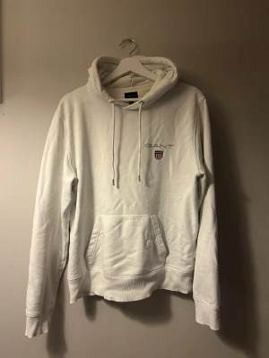Vit hoodie från GANT med logga - Vit hoodie från GANT i storlek L med broderad logga och emblem på bröstet. Tröjan har huva med snörning, känguruficka och ribbade muddar vid ärmslut och nederkant. Perfekt för chill dagar och enkel att matcha med jeans eller joggers. Den är i storlek L men passar mer som M