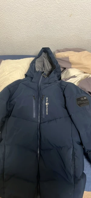 Mörkblå Sail Racing dunjacka - Säljer en mörkblå dunjacka från Sail Racing med huva och två bröstfickor med dragkedja. Jackan har Sail Antarctica-märke på ärmen och Sail Racing-logga på bröstet. Perfekt för kalla dagar med sin tjocka och puffiga design.