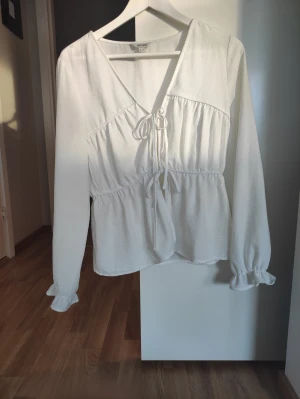 Vit omlottblus med snörning från AWARE - Supersöt vit blus från AWARE i storlek S. Blusen har omlottdesign med v-ringning och snörning framtill, samt långa ärmar med volangdetalj vid ärmslutet. Den är lätt rynkad vid midjan, skitsnygg till Jeans en vanlig dag men kan även kläs upp till en fin middag. 