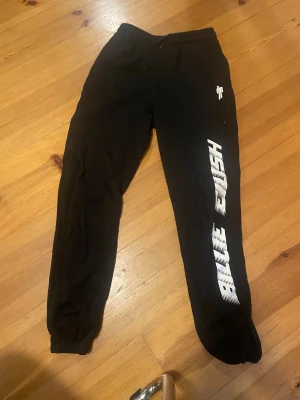 Svarta mjukisbyxor Billie Eilish H&M - Svarta mjukisbyxor från H&M med tryck i vitt längs benet där det står 'Billie Eilish'. Byxorna har resår i midjan och vid bensluten, samt en liten vit symbol på höften. Perfekta för chill eller streetwear.